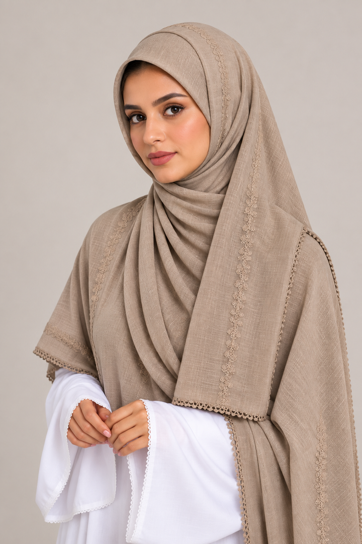 A woman wearing a draped taupe-beige hijab with delicate embroidered borders and a matching shawl draped over her shoulders, paired with a white long-sleeve top, against a solid light gray background.