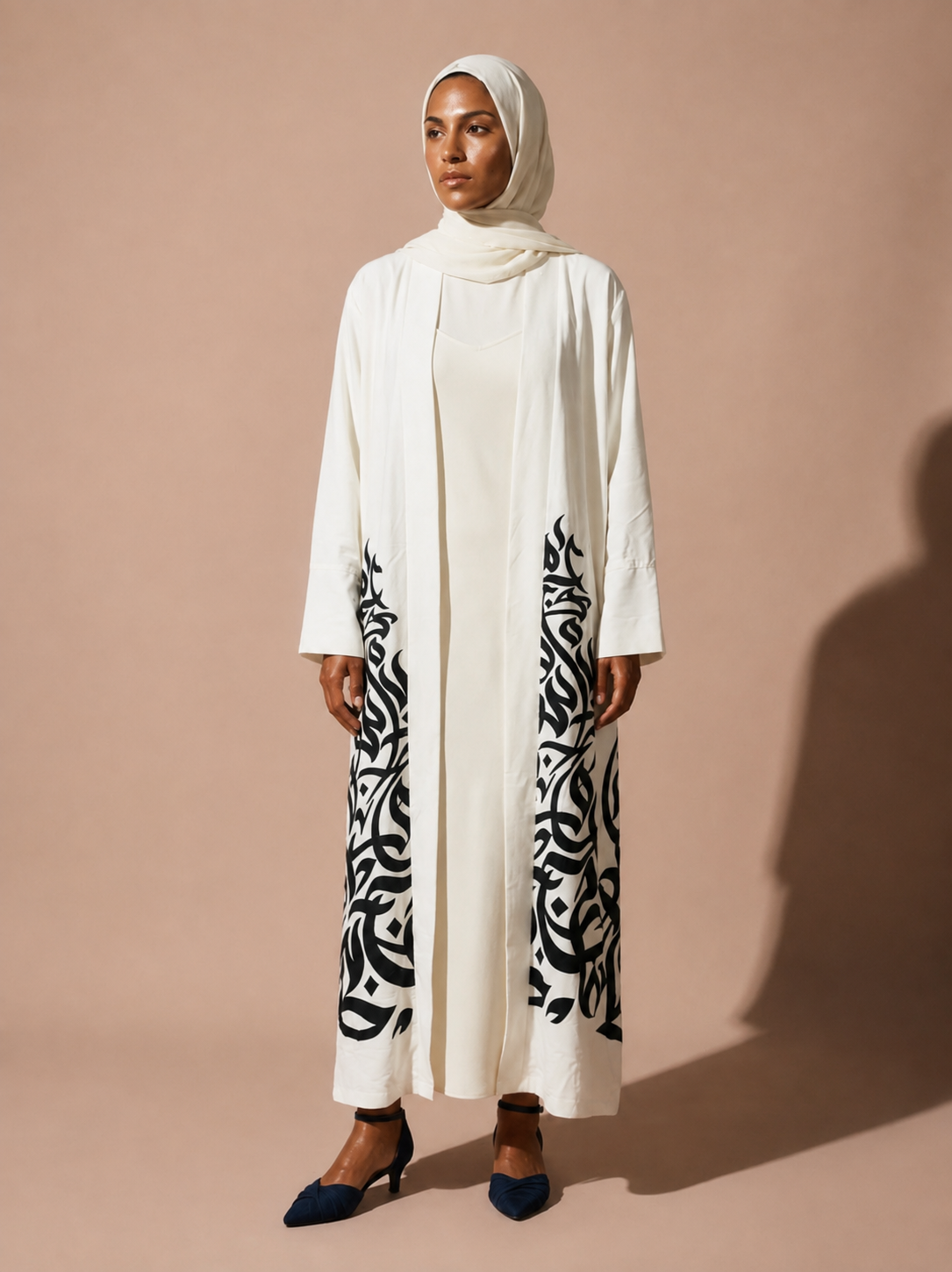A woman wearing a long white abaya with black abstract patterns on the lower front panels, a matching white hijab, and dark blue pointed flats, standing against a solid beige studio backdrop.