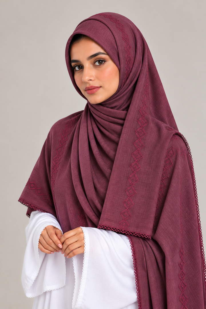 A woman wearing a deep plum-colored embroidered hijab and a matching shawl over a white outfit, photographed against a neutral light gray background.