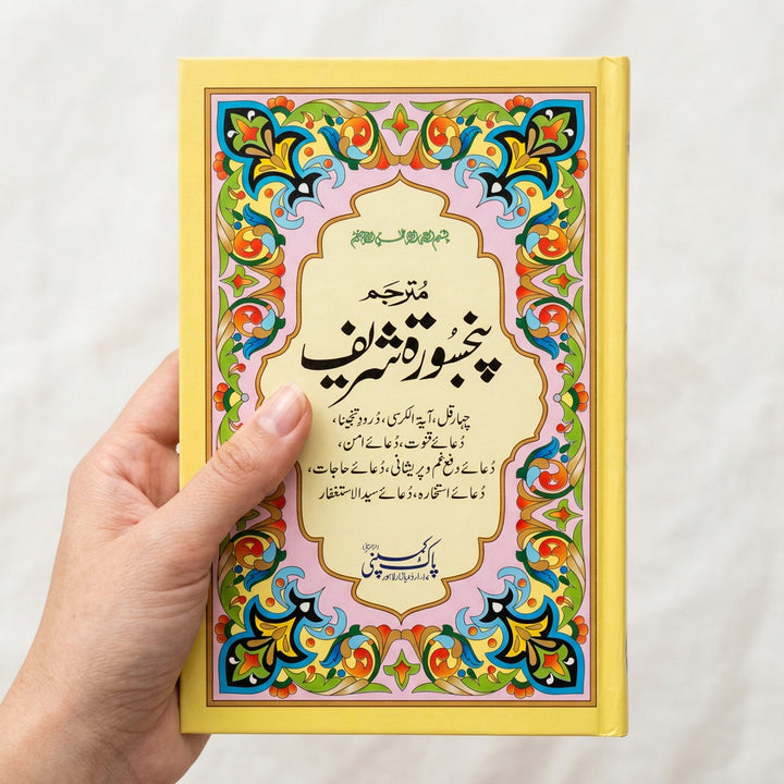 A hand holds a book with a bright yellow cover featuring ornate multicolor Arabic calligraphy and decorative floral borders. The book cover has Urdu text and is photographed in a real indoor setting with shelves and other items visible in the background.