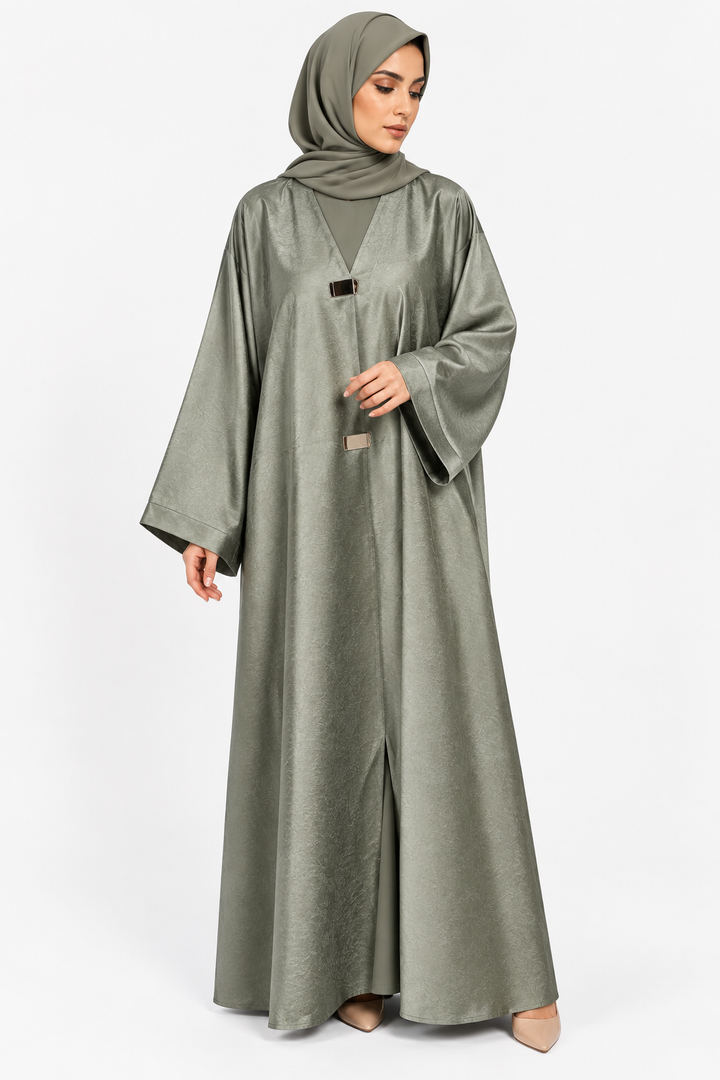 A woman wearing a flowing sage green abaya with a matching hijab, standing against a plain light gray studio background.