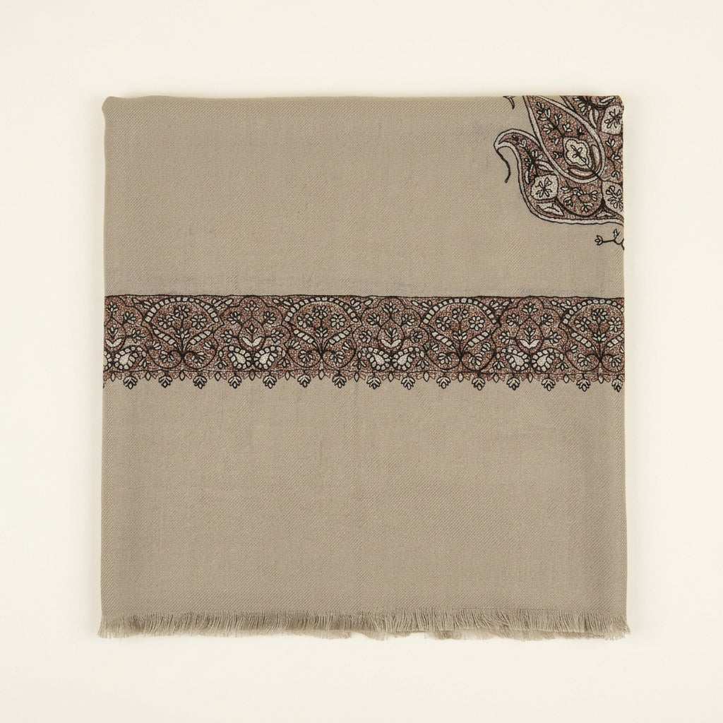 A folded beige fabric piece with a wide embroidered band across the middle and a paisley motif in the upper right corner, placed on a dark tabletop with a menu, white card, and packaged item nearby.