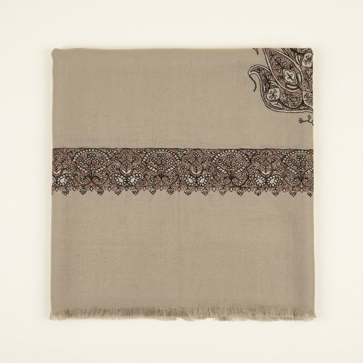 A folded beige fabric piece with a wide embroidered band across the middle and a paisley motif in the upper right corner, placed on a dark tabletop with a menu, white card, and packaged item nearby.