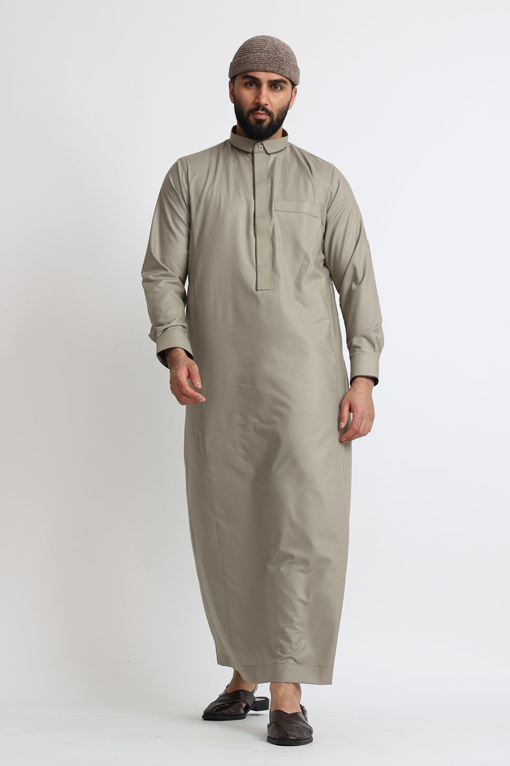 A man wearing a beige thobe with Greek pattern lace trim around the collar and cuffs, along with two side pockets and a left chest pocket.