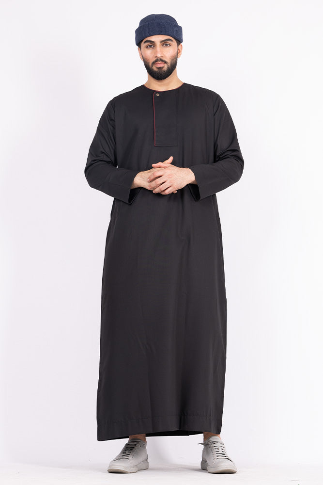 A person standing in a studio setting, wearing a black thobe with maroon piping and a single button detail.